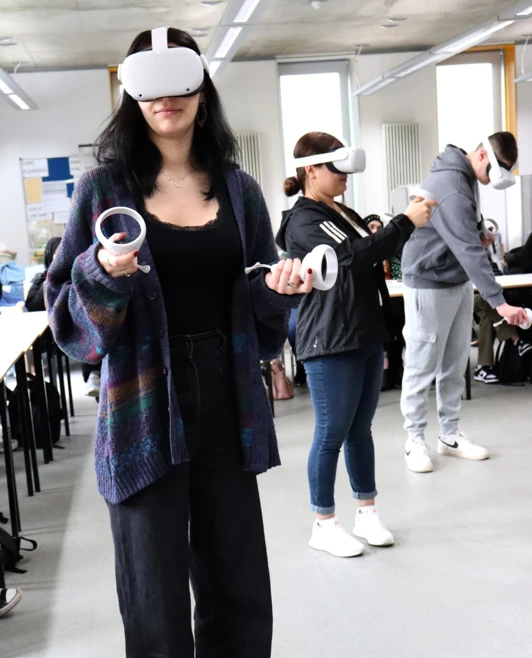 A young woman wearing a VR headset and holding controllers participates in a virtual training session, with other students in the background engaging in the same activity. A young woman wearing a VR headset and holding controllers participates in a virtual training session, with other students in the background engaging in the same activity.